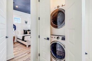A laundry room with two front-loading washing machines and a bed with a white headboard in the adjacent bedroom. - Palmilla Beach Resort-222 Sundew Lane (Port Aransas)