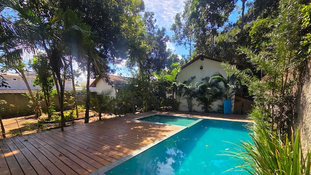 A swimming pool with a wooden deck, surrounded by lush greenery and a building with a tiled roof. - Pousada Luar de Pitinga (Arraial d'Ajuda)