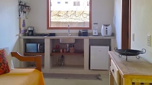 A compact room with a bed, a small desk, a window, and a kitchenette. - Pousada Luar de Pitinga (Arraial d'Ajuda)