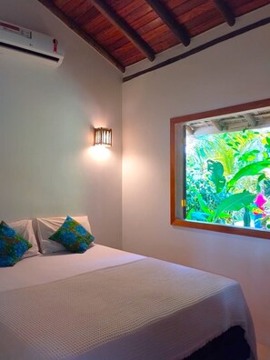 A bedroom with a bed, pillows, a wall-mounted light, an air conditioning unit, and a window with a view of greenery. - Pousada Luar de Pitinga (Porto Seguro)