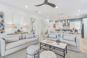 Ein modernes Wohnzimmer mit einer Couch, Hocker und Couchtisch, einer Treppe, die zum Obergeschoss führt, und einer Küchenzeile mit weißen Schränken und Edelstahlgeräten.