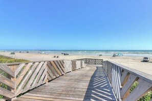 Ein hölzerner Steg führt zu einem Strand, in der Ferne sind Fahrzeuge und Personen zu sehen.