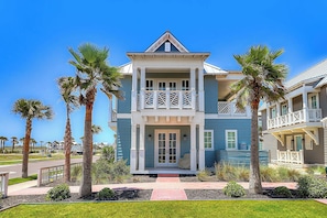 A two-story beachfront house with a balcony, palm trees, and a clear blue sky. - Palmilla Beach Resort-236 Sunset Avenue (Port Aransas)