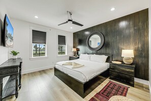 A bedroom with a bed, bedside tables, a ceiling fan, a large mirror, and a view through the window. - Palmilla Beach Resort-244 Sunrise Avenue (Port Aransas)