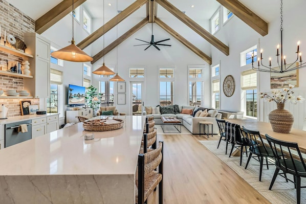 Een ruime woonkamer met een groot centraal eiland, een eetgedeelte met moderne stoelen, en een keuken met een tegelachterwand en houten balken.