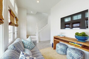 A modern living room with a staircase, a sofa with patterned cushions, a wooden side table with a bowl of fruit, and a wall-mounted TV. - Palmilla Beach Resort-300 Mermaid Tail Lane (Port Aransas)