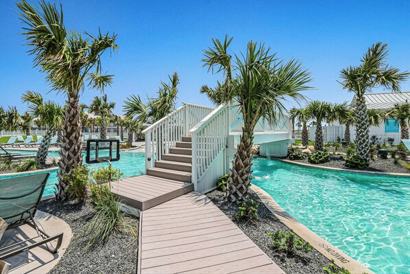 Un área de piscina con muelle de madera, palmeras y una escalera que lleva a un edificio.