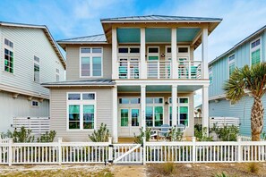Une maison à deux étages située en bord de mer, dotée d’un balcon, d’une clôture blanche et d’un palmier.