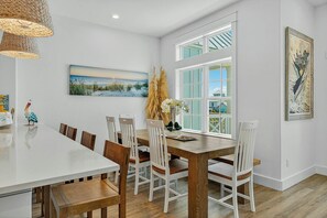A dining area with a white table and chairs, a painting of a beach scene, and a window with a view of houses. - Palmilla Beach Resort-602 Sunset Avenue (Port Aransas)