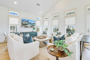 A spacious living room with a high ceiling, large windows, and a view of a pool and buildings. - Palmilla Beach Resort-723 Sunset Avenue (Port Aransas)