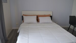 Cama bem arrumada com roupa de cama branca e duas almofadas, parede lisa e uma cadeira ao lado.