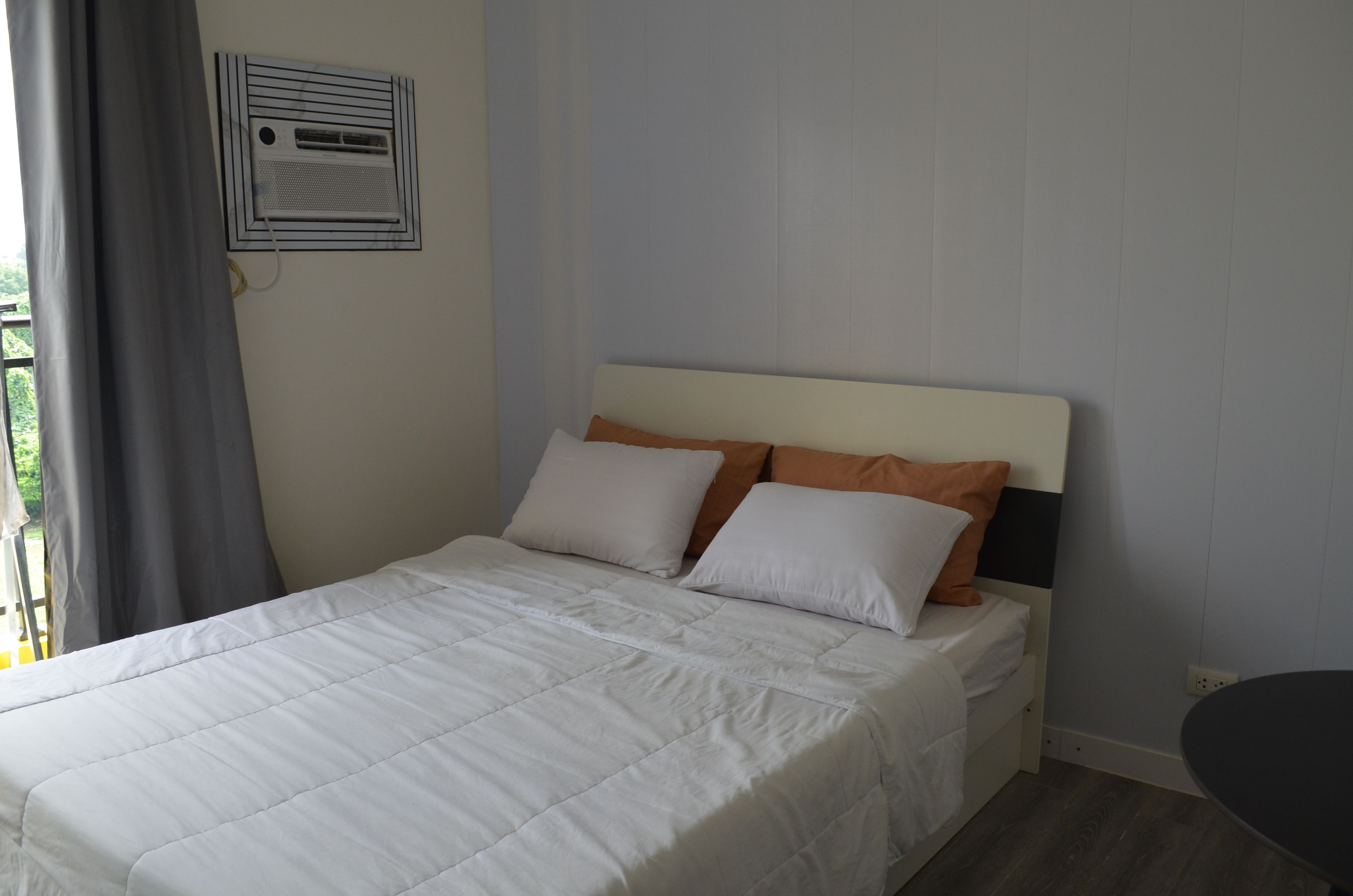 Ein ordentlich bezogenes Bett mit weißer Daunendecke und zwei Kissen, ein Fenster mit Vorhängen und eine Wand-Klimaanlage.