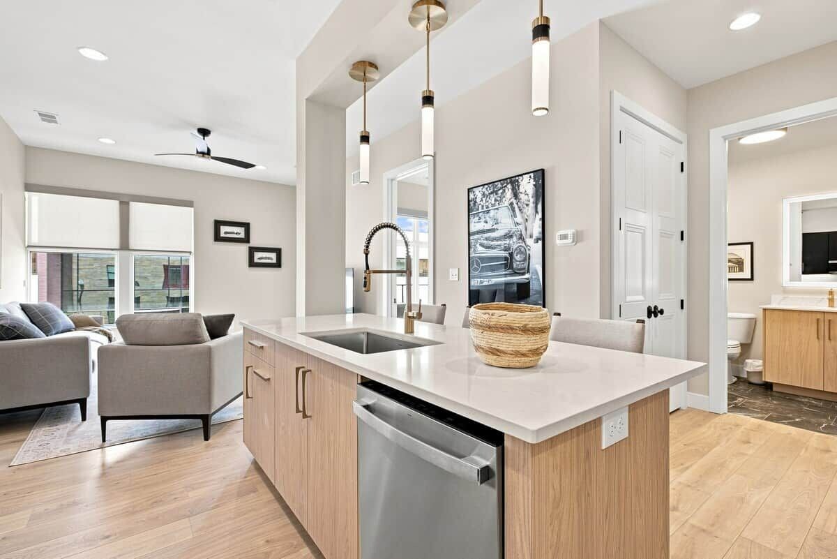 A modern kitchen with a central island, built-in dishwasher, pendant lighting, and a large window.