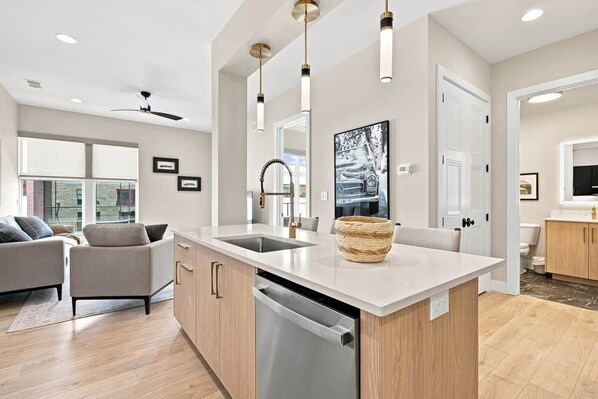 A modern kitchen with a central island, built-in dishwasher, pendant lighting, and a large window.