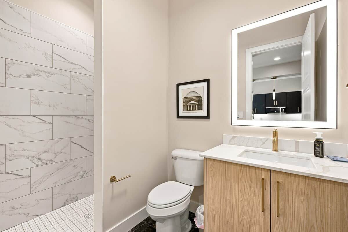 A bathroom with a marble-tiled shower wall, a toilet, a vanity with a mirror, and a framed picture on the wall.
