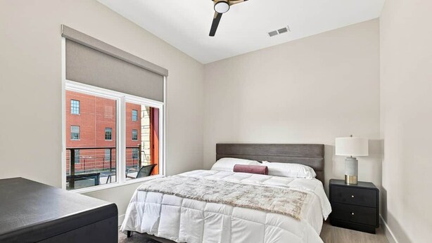 A bedroom with a bed, a nightstand, a lamp, a window with a view of a building, and a ceiling fan. - The Oaks · 2 Bedroom · 2 Bath · 1400SF (Owensboro)