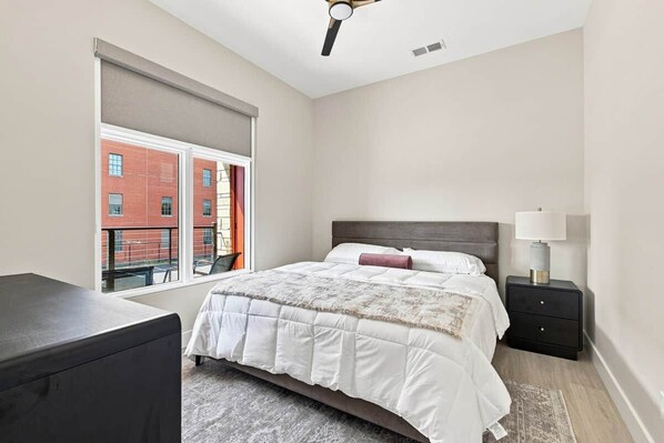 A bedroom with a bed, a nightstand, a lamp, a window with a view of a building, and a ceiling fan.