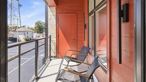 A balcony with two chairs, a glass door, and a view of a parking lot and buildings. - The Oaks · 2 Bedroom · 2 Bath · 1400SF (Owensboro)