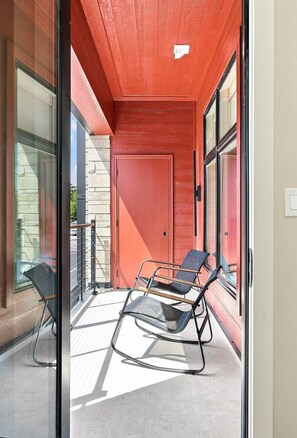 A narrow balcony with a red door, glass railings, and two black chairs.