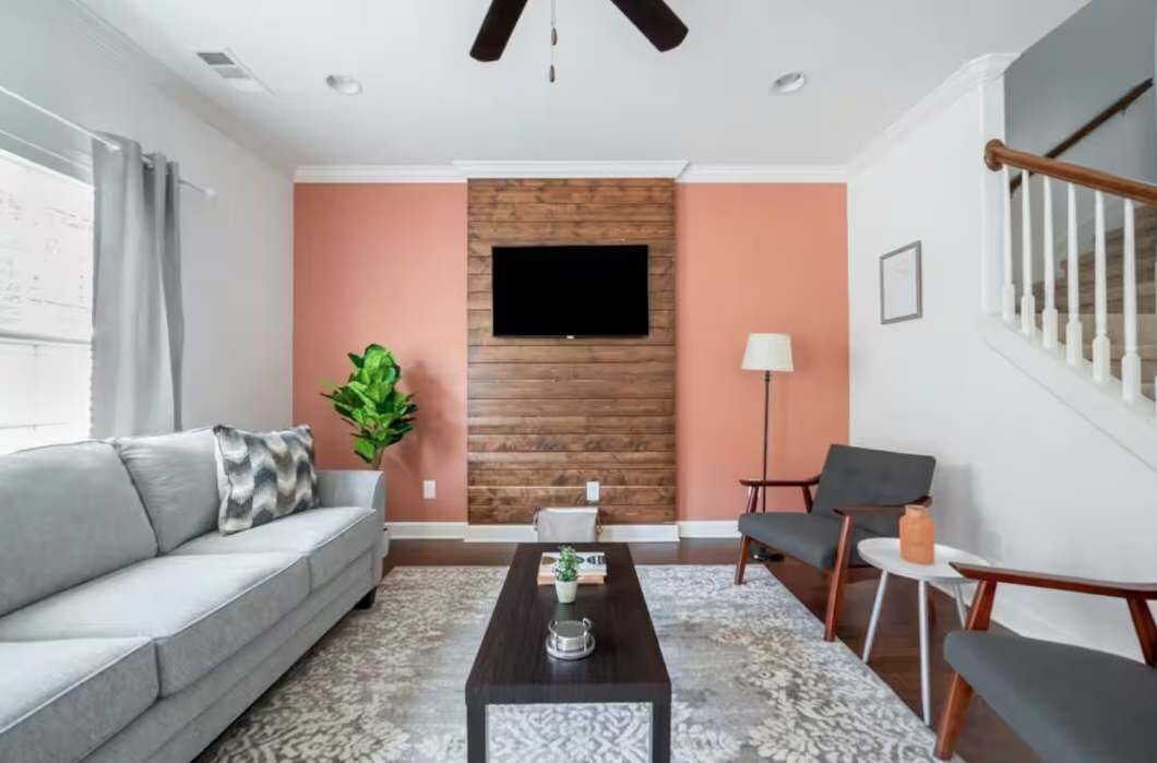 A living room with a sofa, coffee table, and chairs, a TV mounted on a wooden wall, and a staircase with a railing.