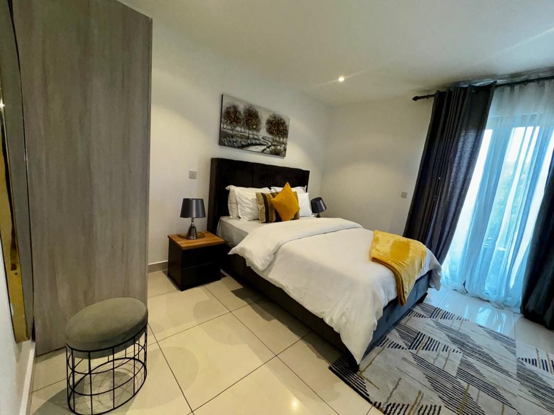A modern hotel room with a large bed, a bedside table with a lamp, a small stool, and a view of the outdoors through a window with curtains.