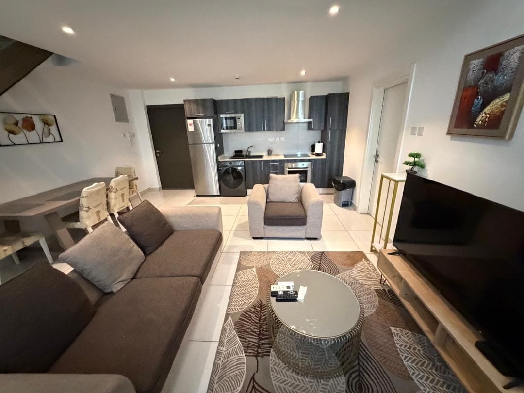 Ett modernt vardagsrum med en soffa, ett soffbord och en tv. I bakgrunden syns ett kök med en mikrovågsugn och en ugn.