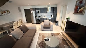 Una sala de estar moderna con un sofá, una mesa de centro y un televisor. Se ve una cocina al fondo, con un microondas y un horno.