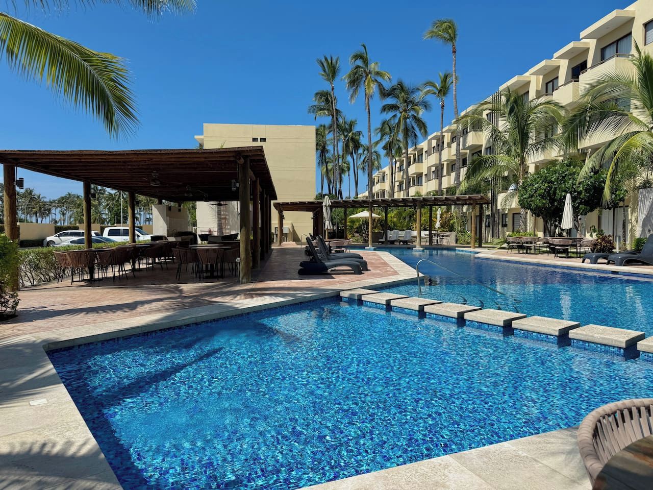 A swimming pool with lounge chairs and a covered seating area, surrounded by palm trees and a multi-story building.