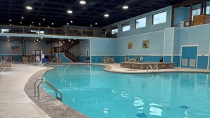 Una piscina interior con superficie de agua cristalina, rodeada por una solana de hormigón y una zona con solana de madera y sillas.