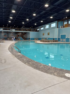 Una piscina interna con superficie d'acqua limpida, circondata da un solarium in cemento e da una zona con pavimento in legno e sedie.