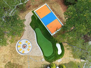 An aerial view of a golf course with a putting green, sand bunkers, and a circular seating area with chairs and a fire pit. - Sleeps 20 | Pickleball/Firepit+Theater/Game Room (Leicester)