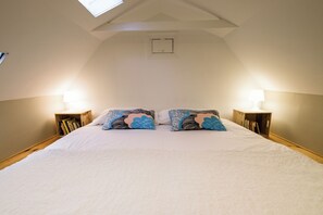 Ein Schlafzimmer mit schrägen Wänden, einem Oberlicht, zwei Nachttischen mit Lampen, einem Bett mit weißer Bettwäsche und einem Bücherregal.