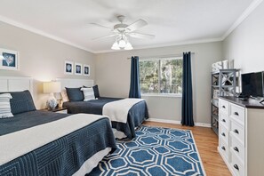 A hotel room with two beds, a ceiling fan, a window with curtains, a desk with a television, and a bookshelf. - Shore Thing- 2 Bed, 2.5 Bath Condo (St. Simons Island)