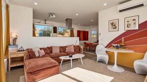 A modern living room with a sofa, coffee table, and chairs. There is a dining area with a table and chairs. The room has large windows and artwork on the walls.