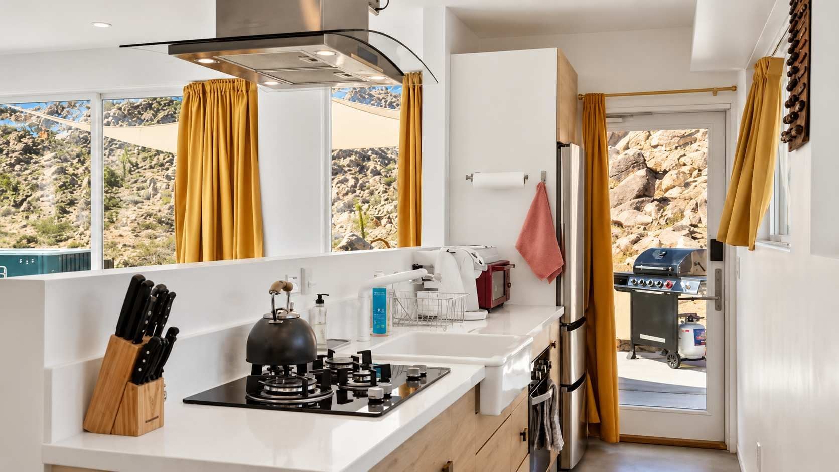 Une cuisine compacte équipée d’une cuisinière à gaz, d’une bouilloire et d’une machine à café. On y trouve une fenêtre avec des rideaux, un évier et une vue sur un paysage rocheux.