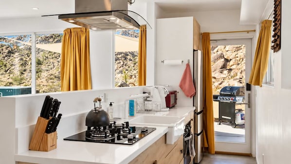 A compact kitchen with a gas stove, a kettle, and a coffee maker. There is a window with curtains, a sink, and a view of a rocky landscape.