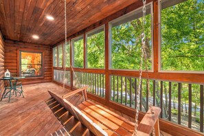 A wooden cabin with a porch, a swing, and large windows overlooking a forest.