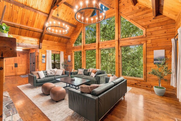 A spacious living area with a large sofa set, a coffee table, and a fireplace. There are multiple windows providing natural light and views of the surrounding greenery.