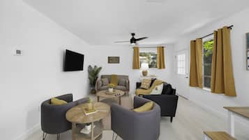 A modern living room with a flat-screen TV, a ceiling fan, and a large window with curtains.