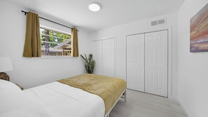Ein Schlafzimmer mit einem Bett, einem Fenster mit Vorhängen, einer Pflanze und weißen Türen.