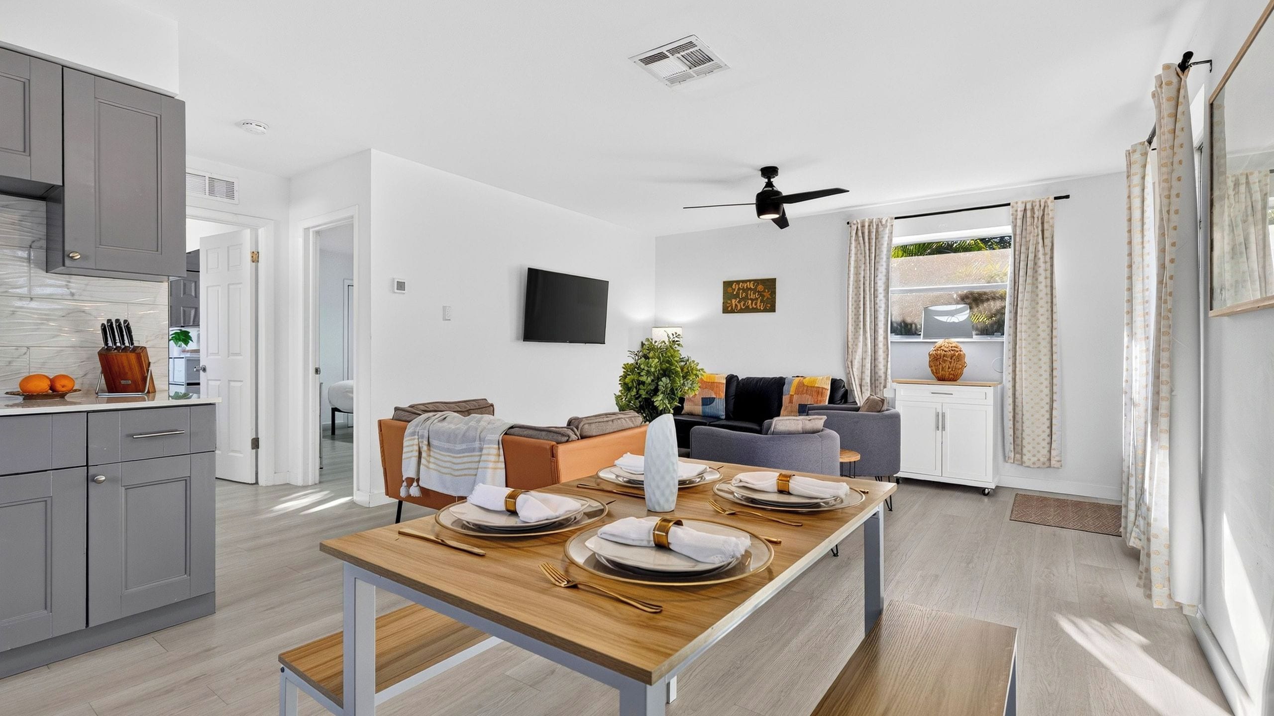 A modern kitchen with grey cabinets, a dining area with a wooden table set for four, and a living room with a sofa and a TV mounted on the wall.