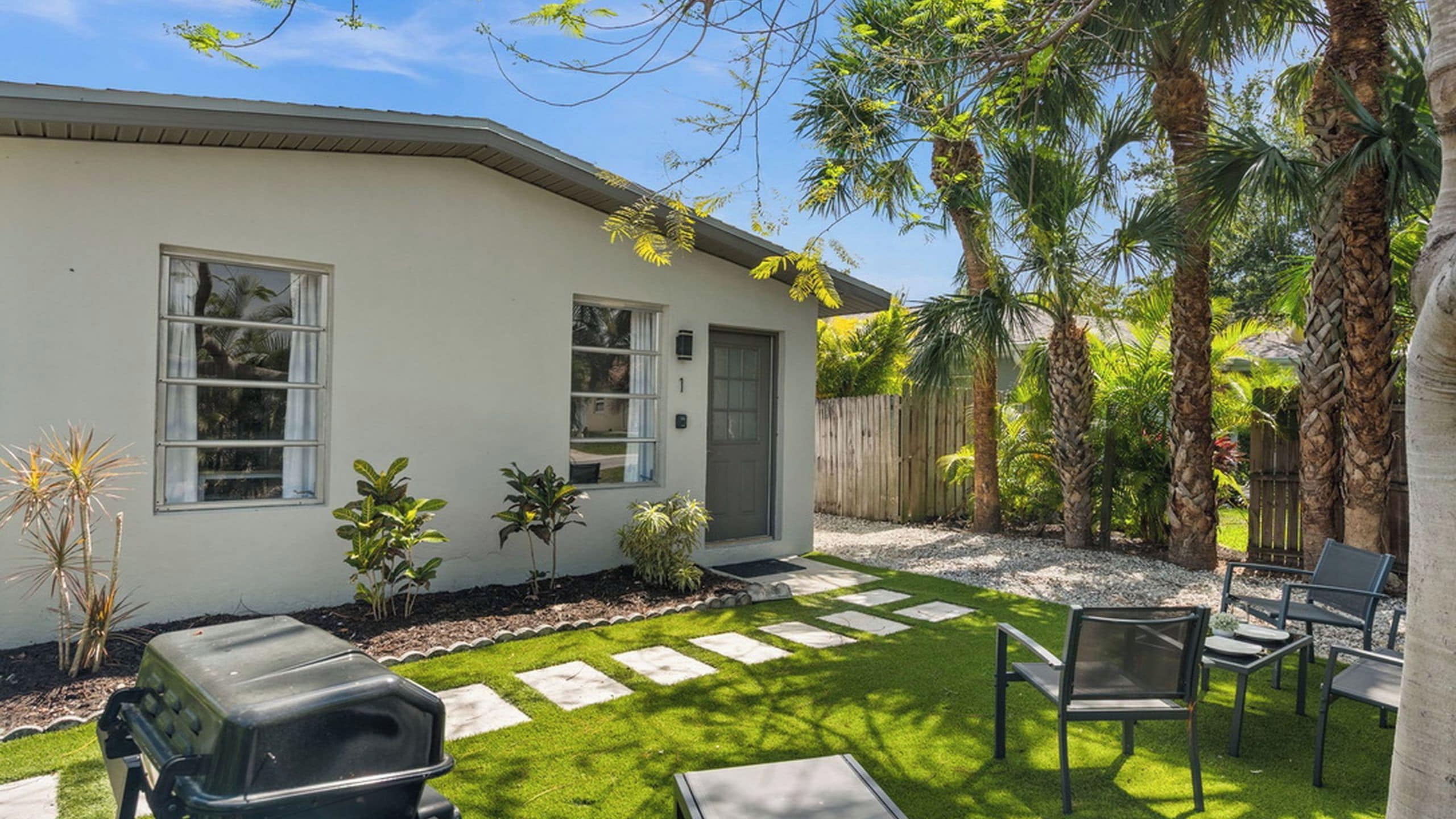 Un immobile residenziale con facciata bianca, una porta d'ingresso, un piccolo giardino con prato e pietre per il passaggio, e un patio con mobili da esterno.