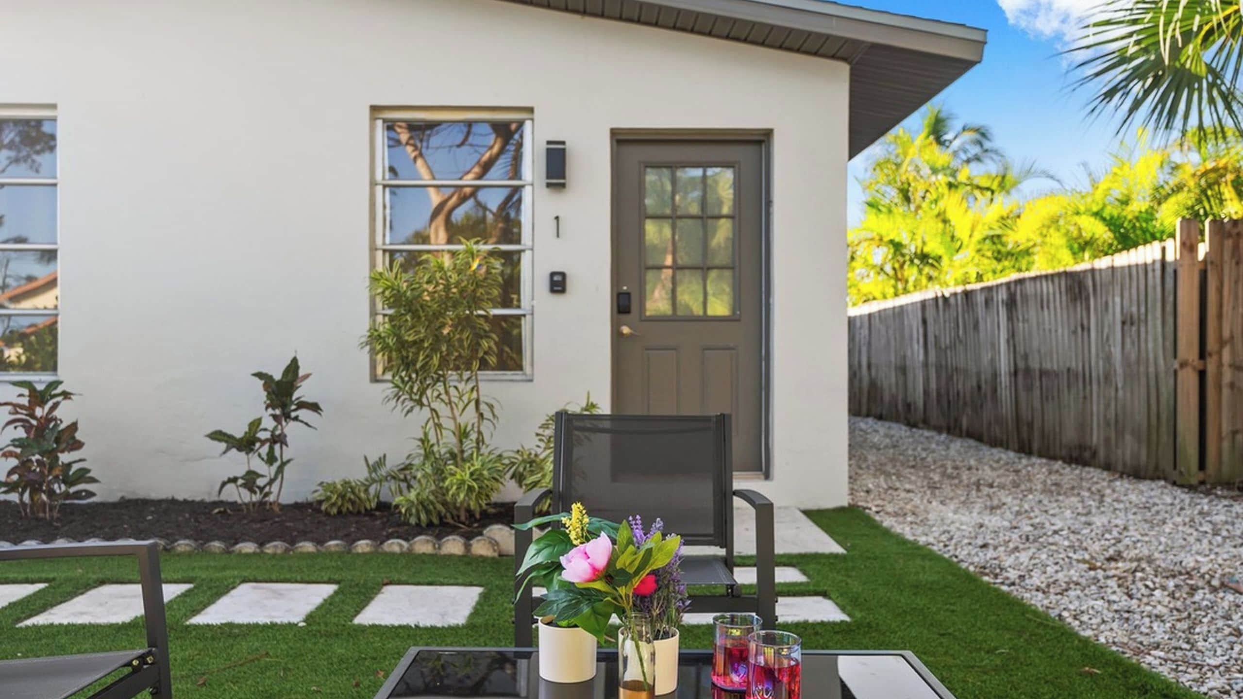 Ein Wohnhaus mit Haustür, einer kleinen Veranda mit Stuhl, einem Tisch mit Blumen in einer Vase und einem Holzzaun.