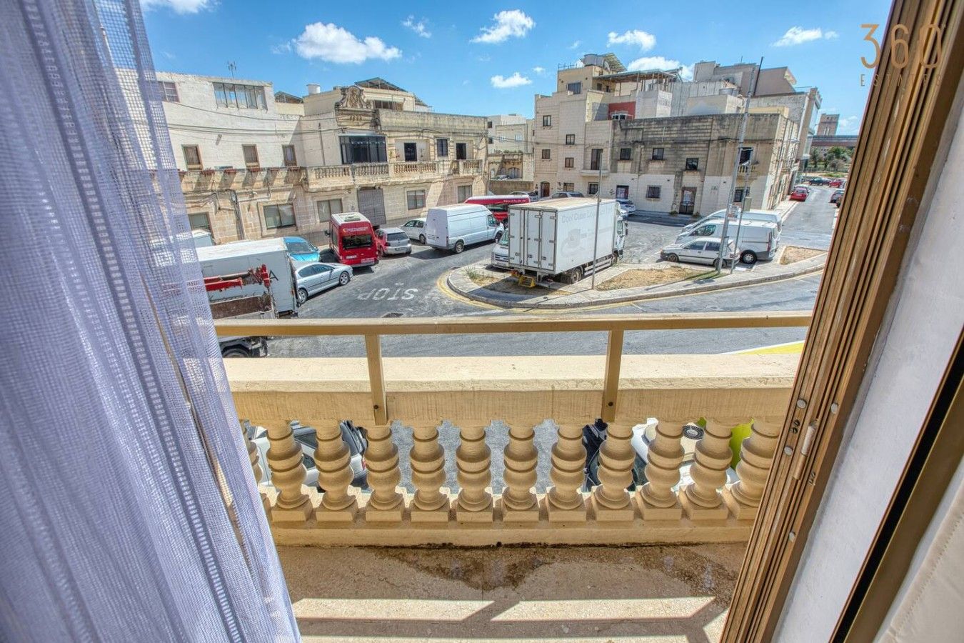 Udsigt fra en balkon over en gade med parkerede køretøjer og bygninger.