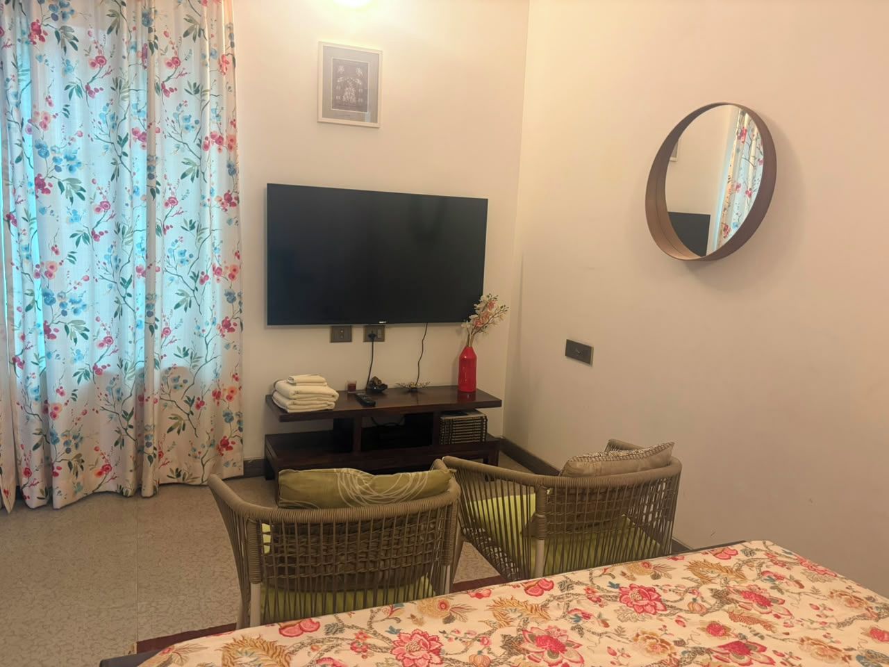 Ein Zimmer mit einem an der Wand befestigten Fernseher, einem Esstisch mit Stühlen, einem Korbsessel, einem kleinen Tisch mit einer Vase und einem blumigen Vorhang.