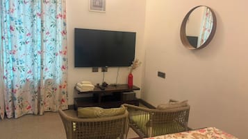 Ein Zimmer mit einem an der Wand befestigten Fernseher, einem Esstisch mit Stühlen, einem Korbsessel, einem kleinen Tisch mit einer Vase und einem blumigen Vorhang.