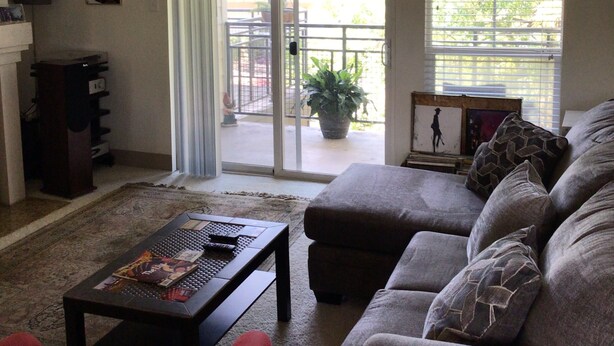 Ein Wohnzimmer mit Deckenventilator, einer Schiebetür zu einem Balkon, einer Couch, einem Couchtisch und einem Fernseher auf einem Standfuß.