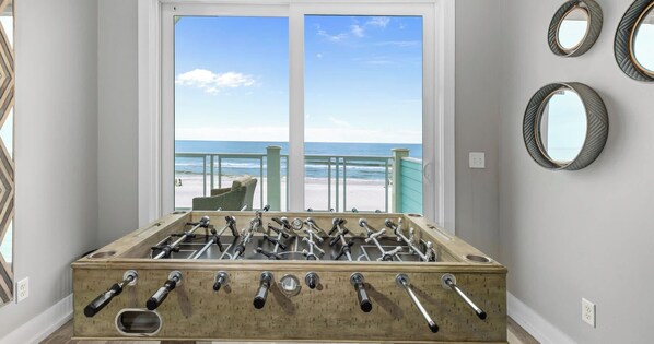 A room with a foosball table, a window with a view of the beach, and three wall-mounted mirrors.