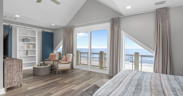 A bedroom with a large window, a bed, a chair, a small table, and a shelf with decorative items.