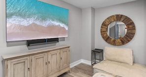 A living room with a large flat-screen TV, a wooden cabinet, a round mirror, a small side table, and a beige sofa.